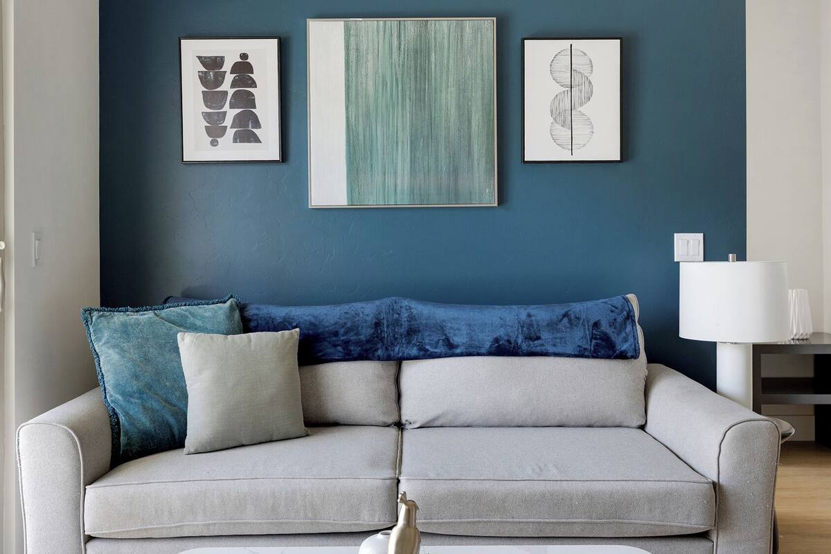 Un salon moderne avec un canapé gris, des coussins bleus et blancs, et trois œuvres d’art encadrées accrochées au mur.