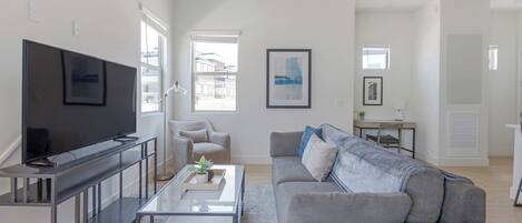 A modern living room with a flat-screen TV, a grey sofa, a coffee table, and a desk with a chair.