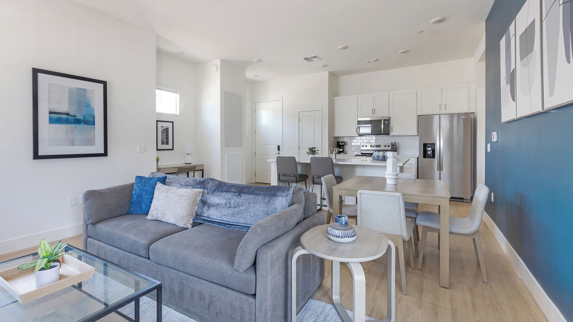 Een modern appartement met een grijze bank, een glazen salontafel, een eethoek met een witte tafel en stoelen, en een keuken met witte kasten en apparatuur.