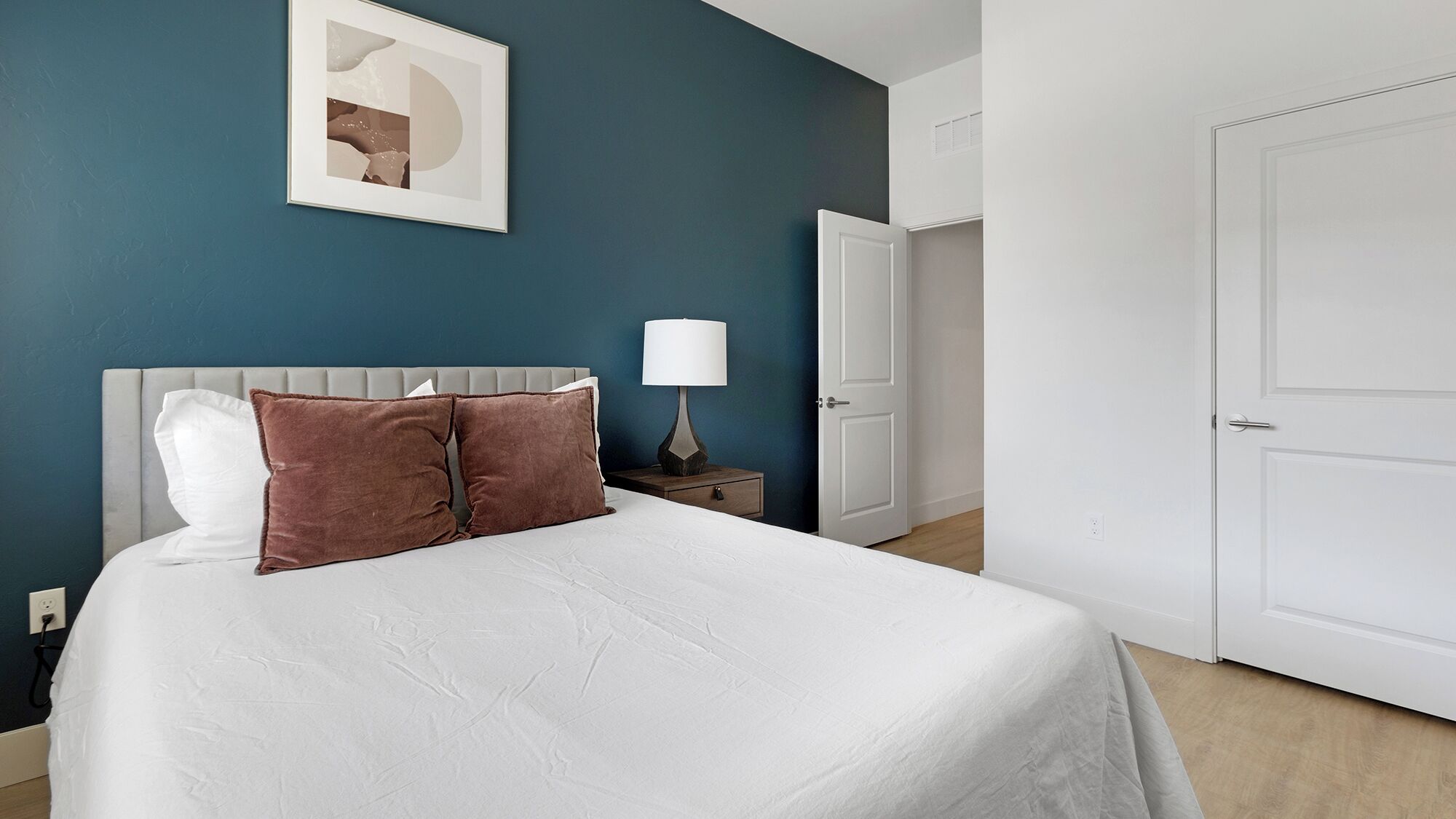 Een slaapkamer met een bed, twee kussens, een nachtkastje met een lamp, een aan de muur hangend schilderij en een deur.