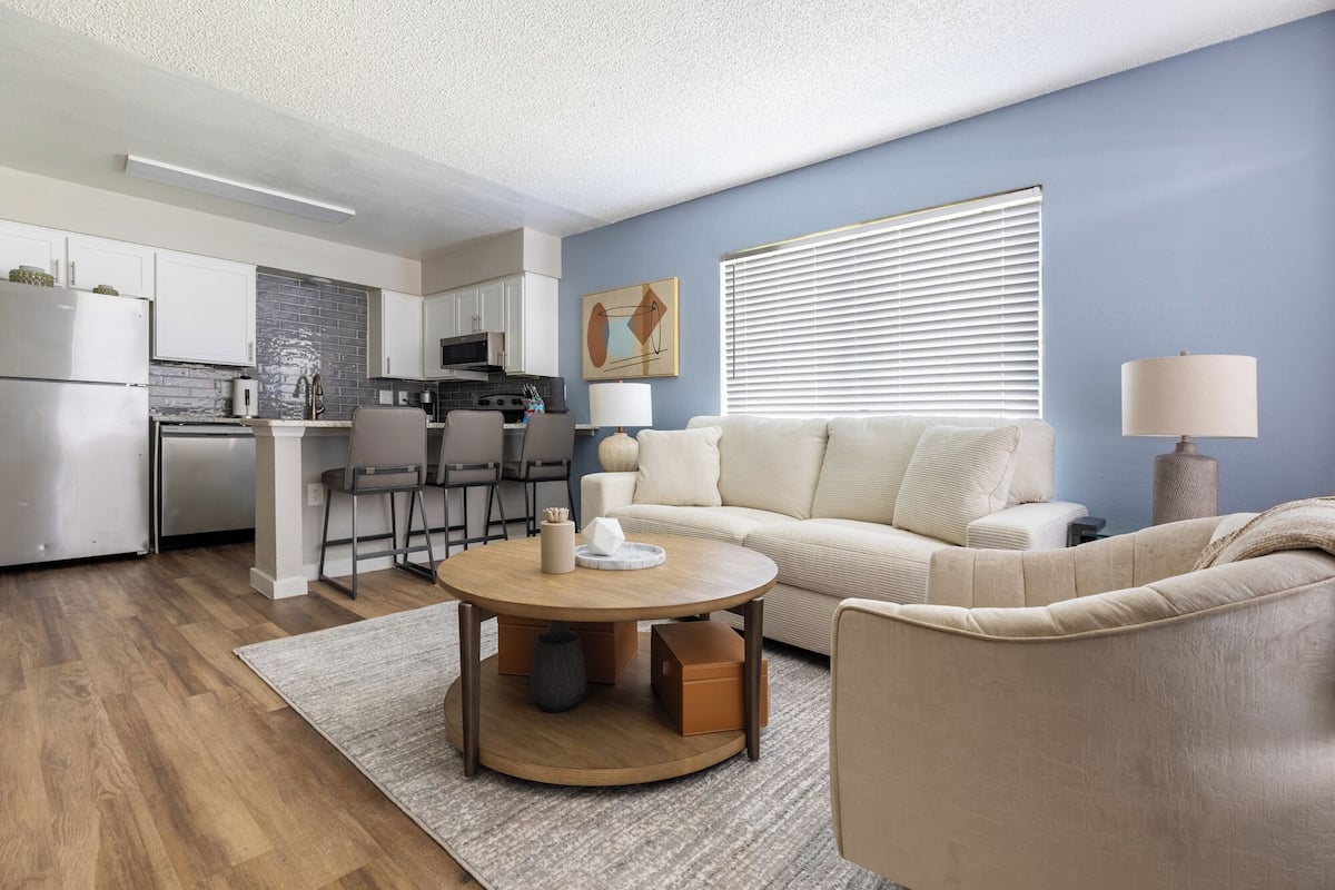 Una sala de estar moderna con un sofá color beige, una mesa de centro redonda y una zona de cocina con armarios blancos y salpicadero gris.