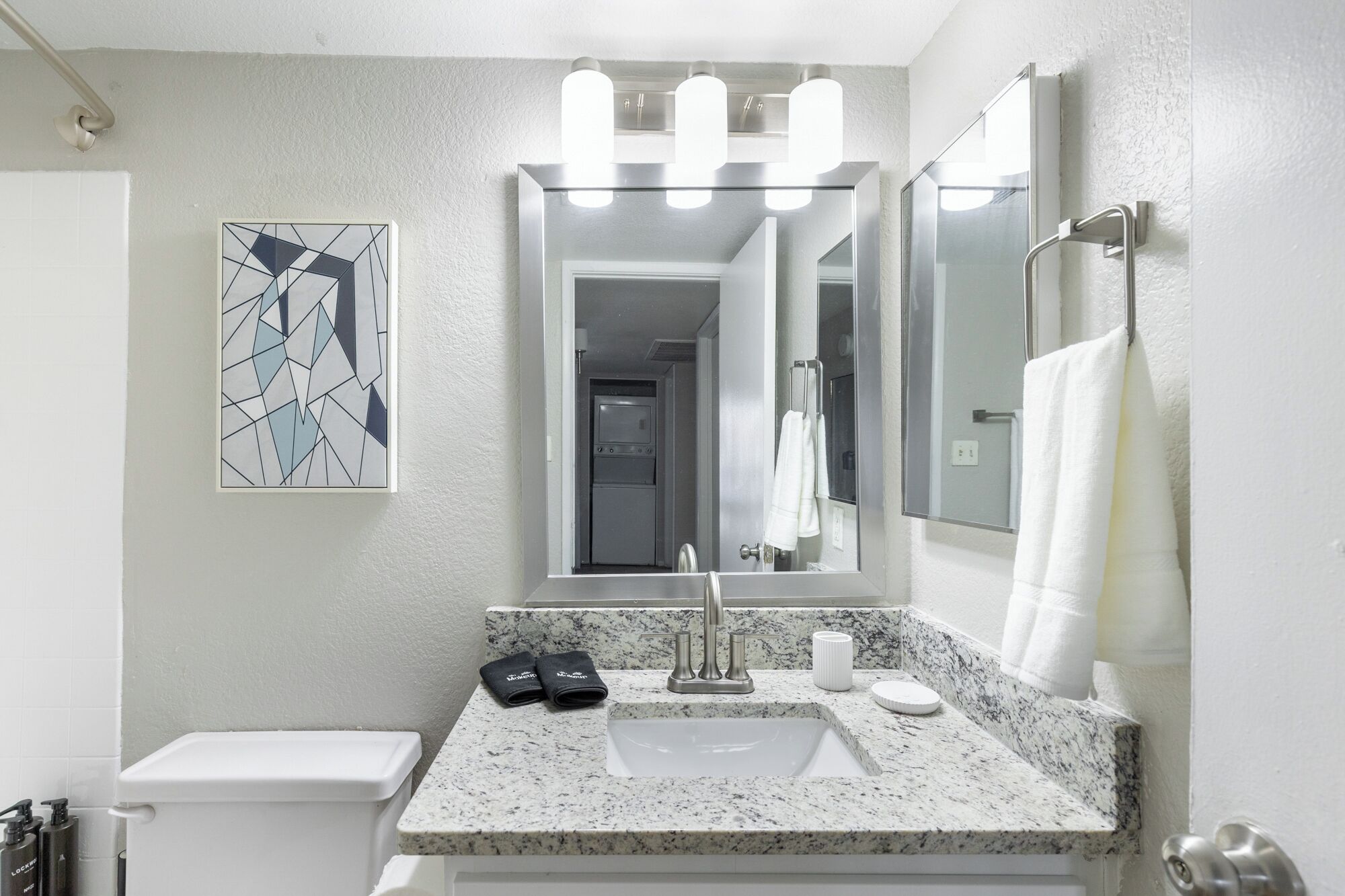 A modern bathroom with a granite countertop sink, a large mirror, and a geometric art piece on the wall.