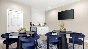 Un espace repas moderne avec une table ronde et des chaises bleues, une cuisine en arrière-plan et un téléviseur à écran plat fixé au mur.