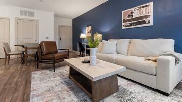 Un salon avec un canapé, une table basse et un fauteuil. Il y a un tableau au mur et une plante sur la table basse.