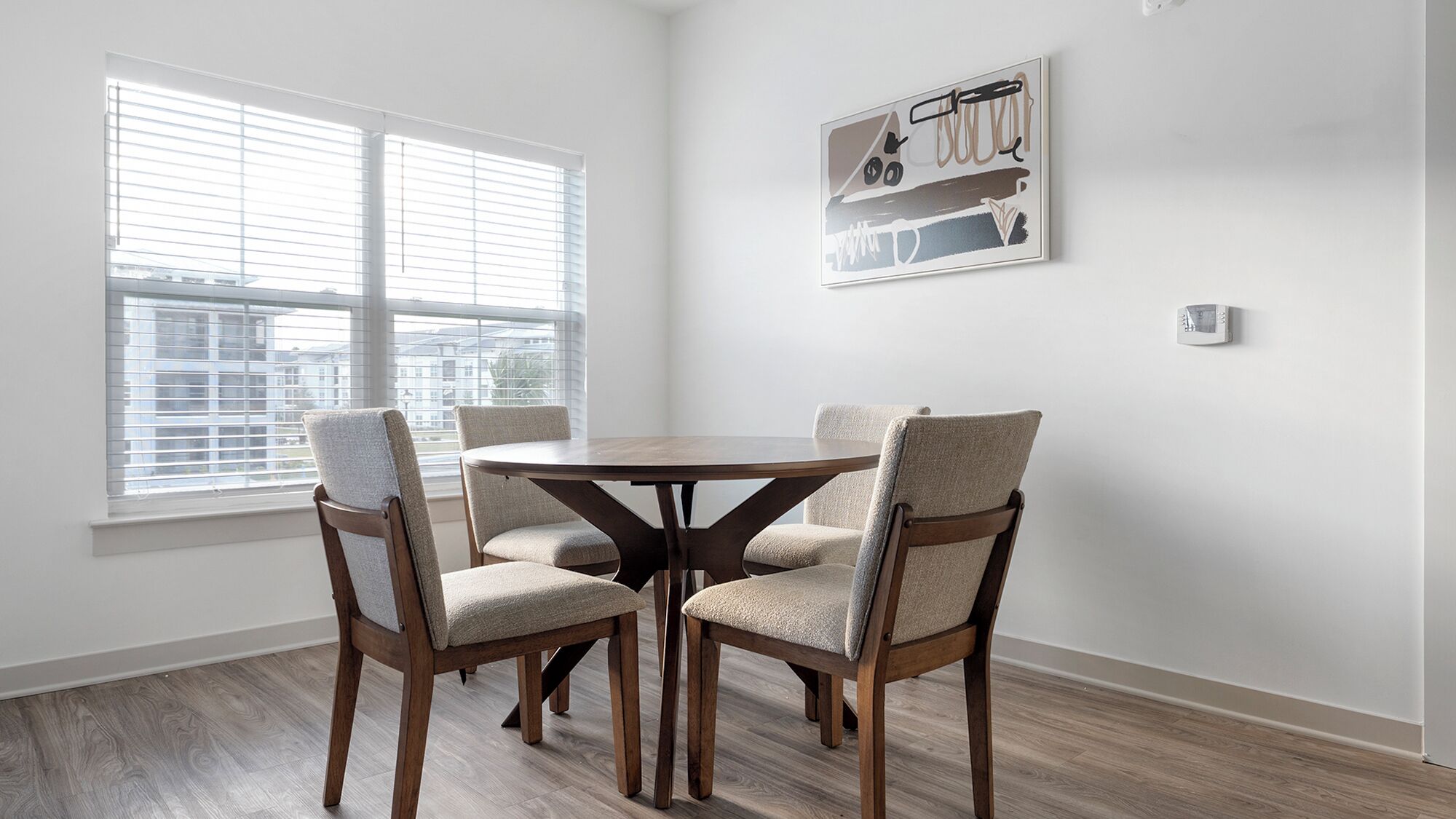A dining area with a round wooden table and four chairs, a window with blinds, and a wall-mounted artwork.