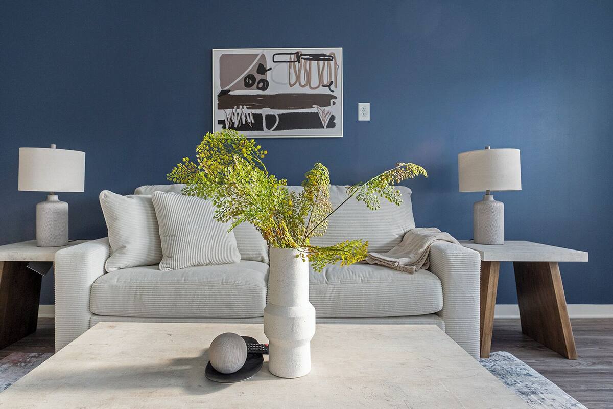 A living room with a blue wall, a grey sofa, two side tables with lamps, a white rug, and a vase with green foliage.