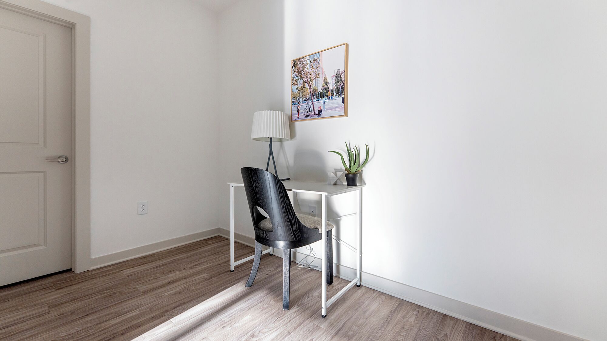 Una habitación con una puerta blanca, un escritorio blanco con una silla negra, una planta en maceta y un cuadro en la pared.