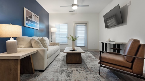 A modern living room with a sofa, coffee table, and a chair. There is a TV mounted on the wall and a ceiling fan. - Landing Apartments - Waterway Hills (Longs)