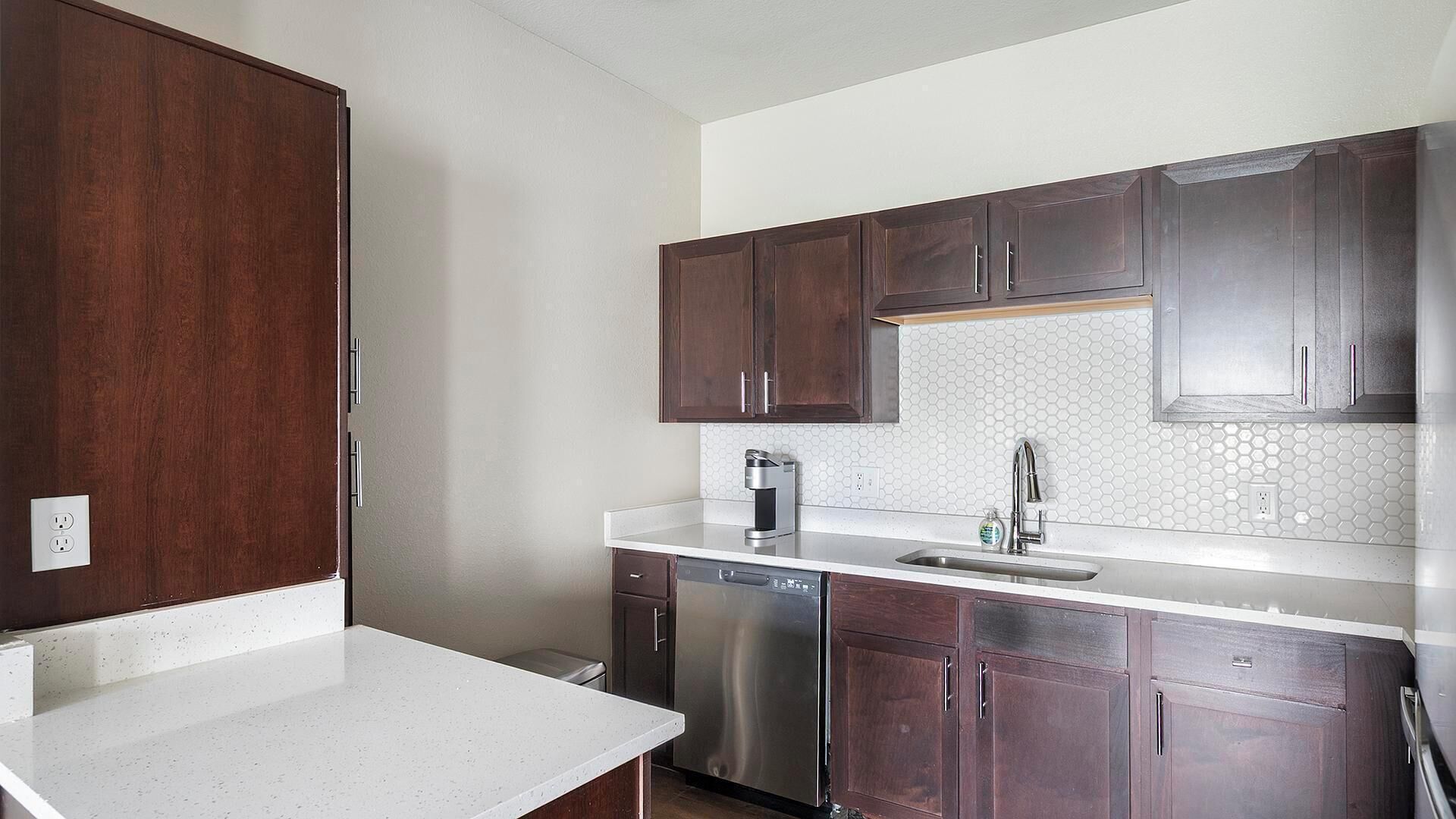 Una cocina con armarios de madera oscura, lavavajillas de acero inoxidable y encimera blanca.