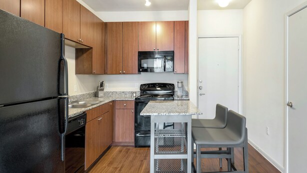A compact kitchen with wooden cabinets, a black refrigerator, a microwave, a stove, and a small dining area with a chair. - Landing Apartments Murfreesboro near Fairfield Estates (Murfreesboro)