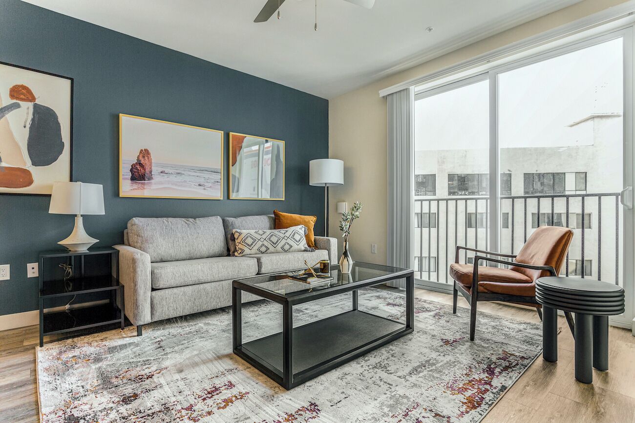 A modern living room with a grey sofa, a glass coffee table, and a leather armchair. There are two framed artworks on the wall and a floor lamp.
