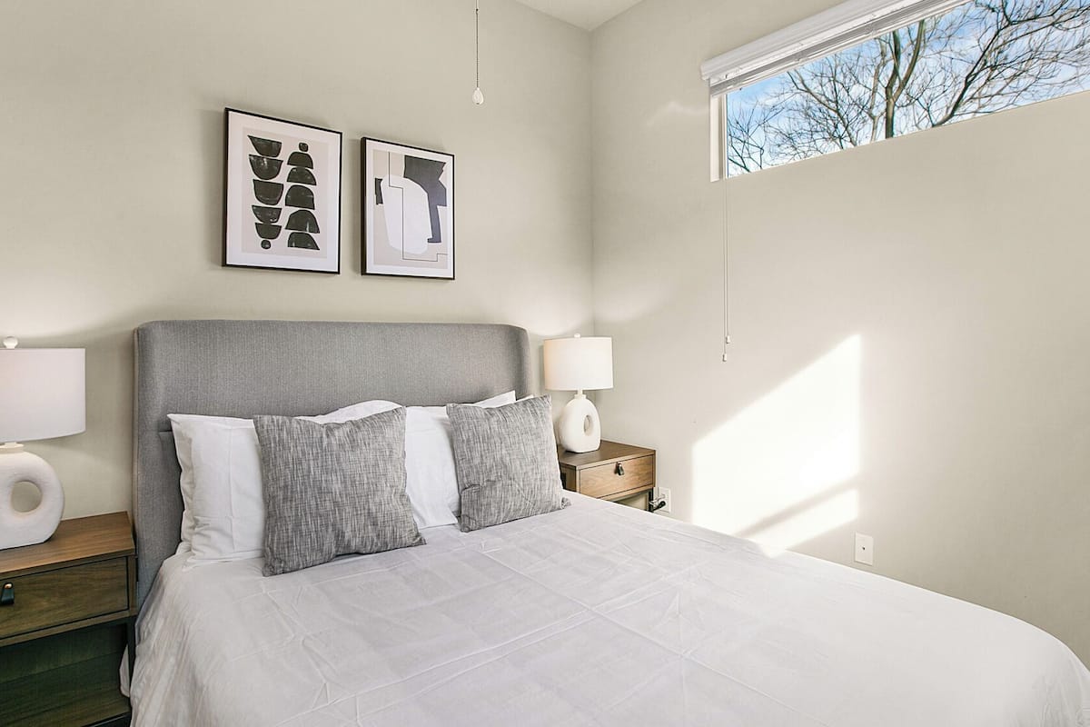 A bedroom with a bed, two bedside tables, a window, and two framed artworks on the wall.