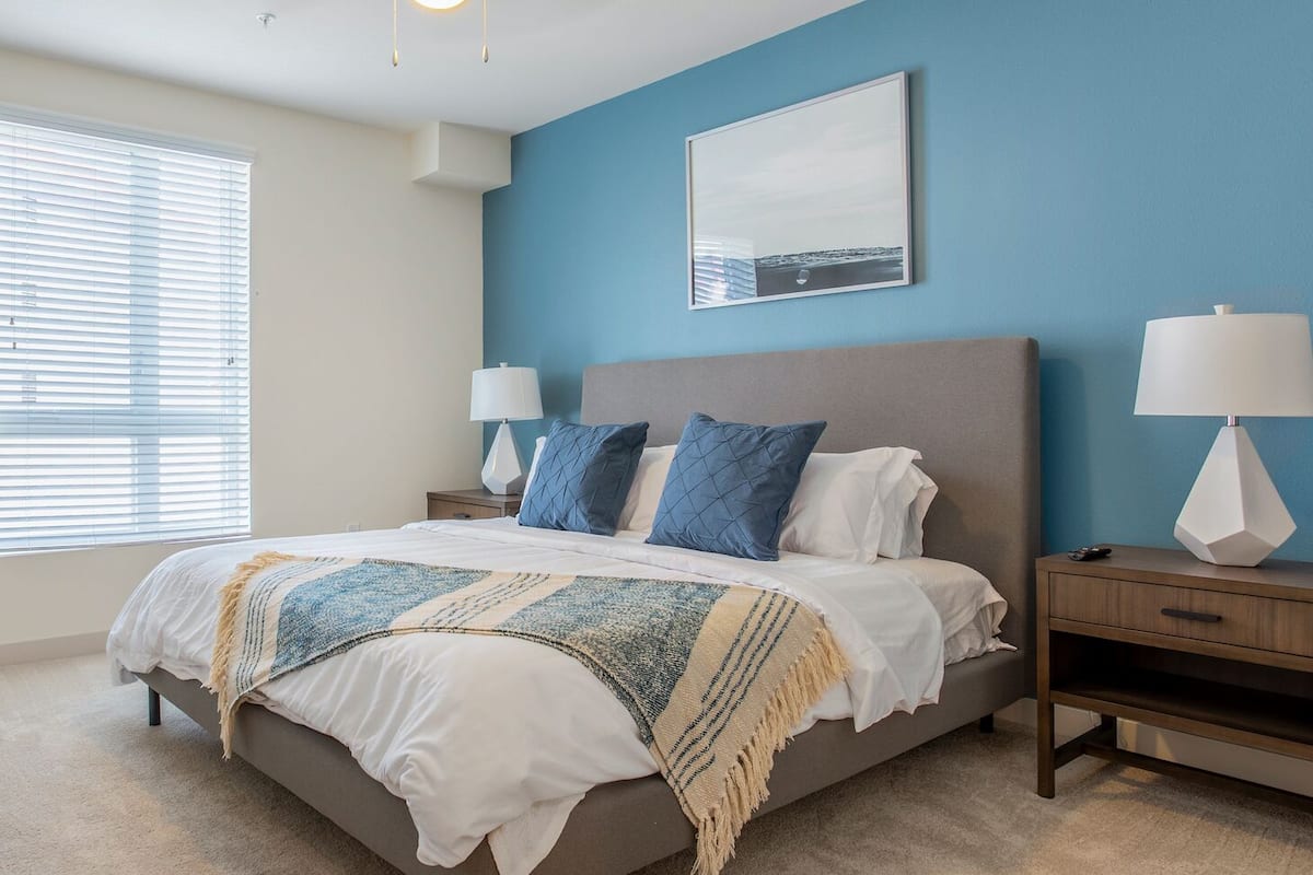 A bedroom with a large bed, two bedside tables, a nightstand, a wall-mounted lamp, and a framed picture.