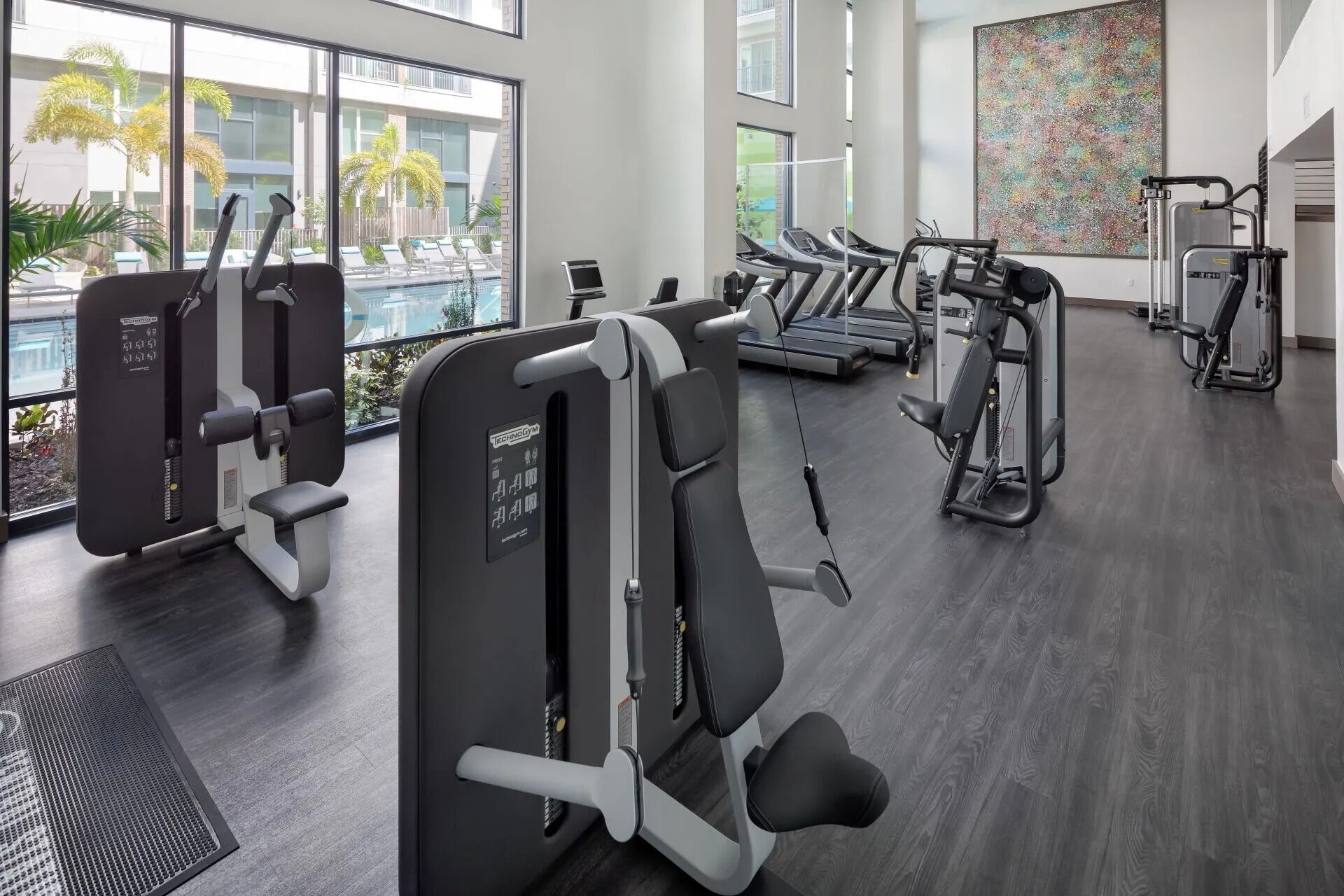 Une salle de sport moderne équipée de divers appareils d’exercice, dotée de grandes fenêtres et offrant une vue sur une piscine et des bâtiments.