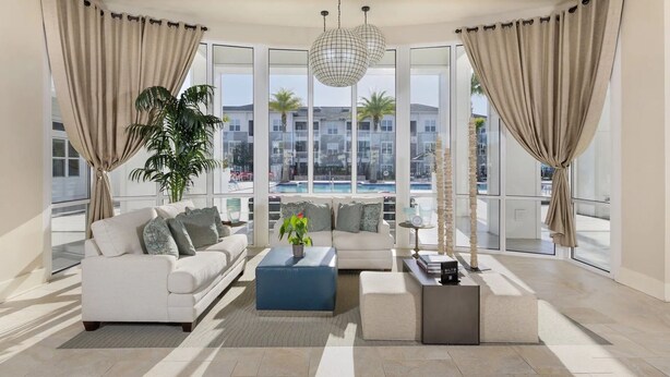 Ein geräumiges Wohnzimmer mit einem weißen Sofa, einem blauen Couchtisch und einer Zimmerpflanze. Große Fenster bieten Blick auf einen Swimmingpool und Gebäude.