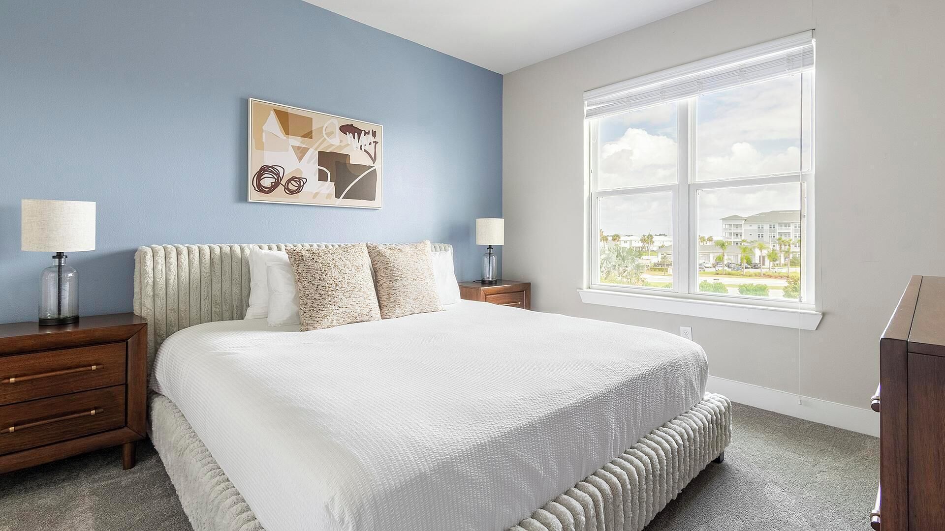 A bedroom with a large bed, two bedside tables with lamps, a window with a view, and a wall-mounted abstract painting.