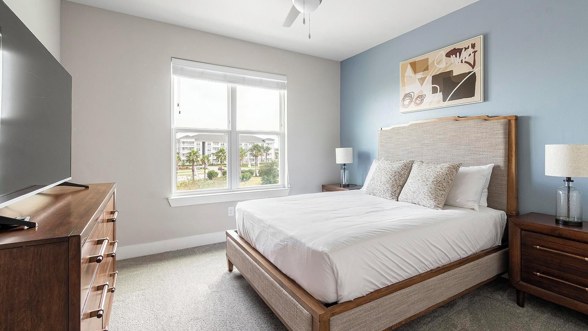 A bedroom with a bed, a TV, a window, and a painting on the wall.