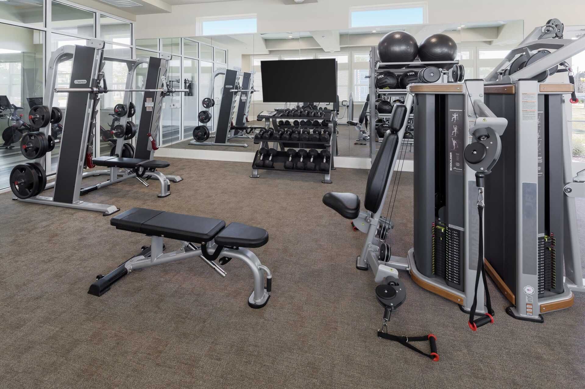 Un gimnasio con equipo de ejercicio variado, incluyendo máquinas de pesas, mancuernas y una banca.