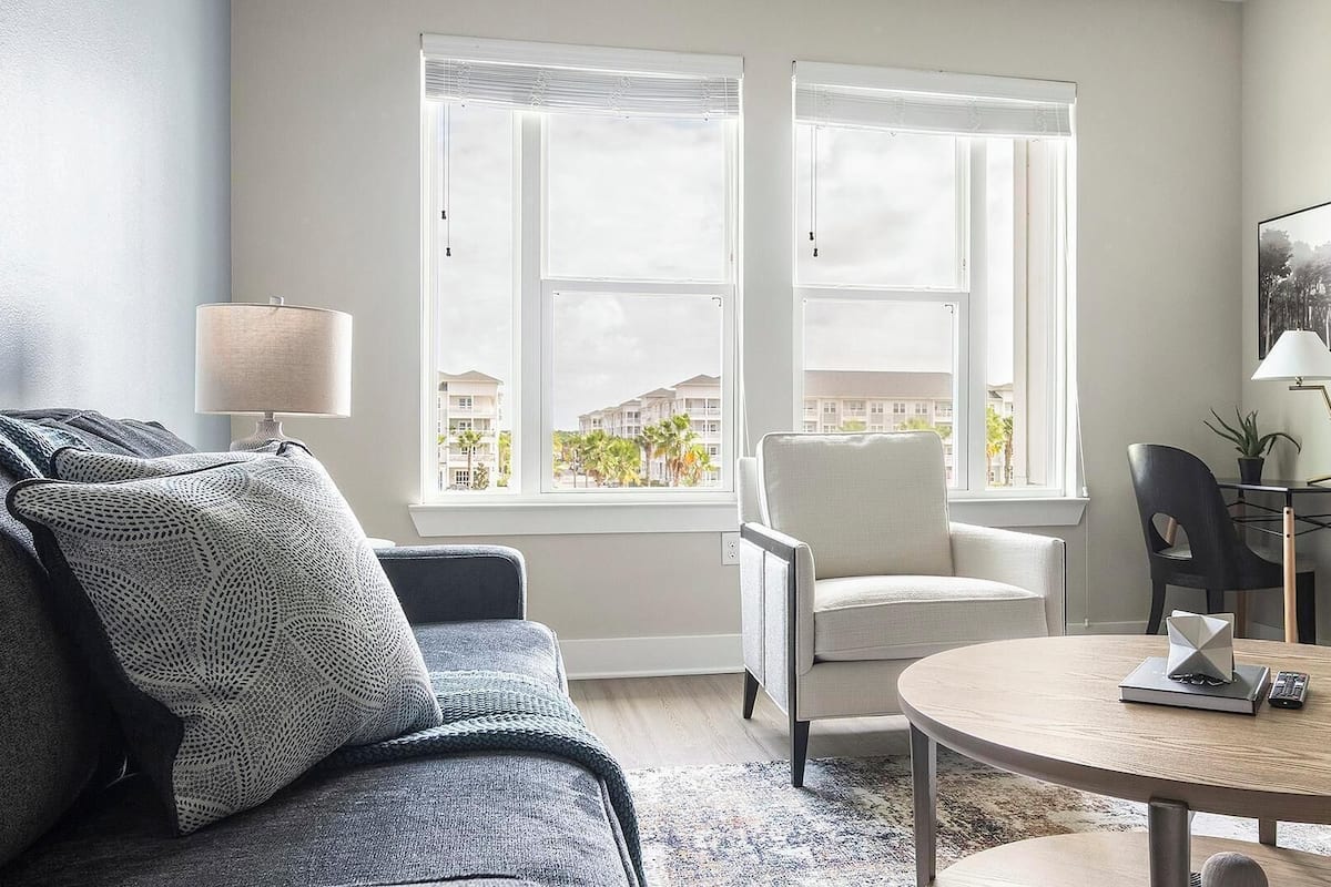 Ein modernes Wohnzimmer mit einem grauen Sofa, einem runden Couchtisch aus Holz und einem weißen Sessel. Zwei Fenster mit Jalousien, eine Lampe auf einem Beistelltisch und ein gerahmtes Bild an der Wand.