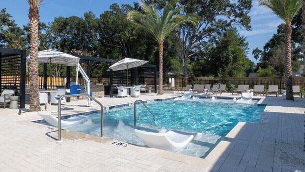 A swimming pool with lounge chairs and umbrellas, surrounded by palm trees and a fence.