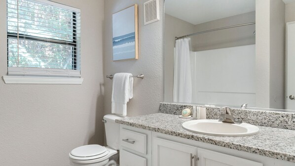 Une salle de bain avec des toilettes blanches, un lavabo sur un plan de travail en granit, un miroir et une fenêtre avec des stores.