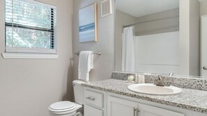 Un baño con inodoro blanco, lavamanos sobre encimera de granito, espejo y ventana con persianas.