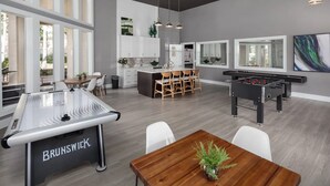 A modern kitchen with white cabinets, a dining area with a wooden table and chairs, and a foosball table. - Landing Apartments Jacksonville - Windy Hill (Jacksonville)