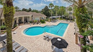 A swimming pool with lounge chairs and umbrellas, surrounded by palm trees and a tiled patio area. - Landing Apartments Jacksonville - Windy Hill (Jacksonville)