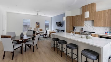 Una cocina moderna con encimera de mármol, armarios de madera y estufa integrada. Hay taburetes de bar y una zona de comedor con mesa y sillas.