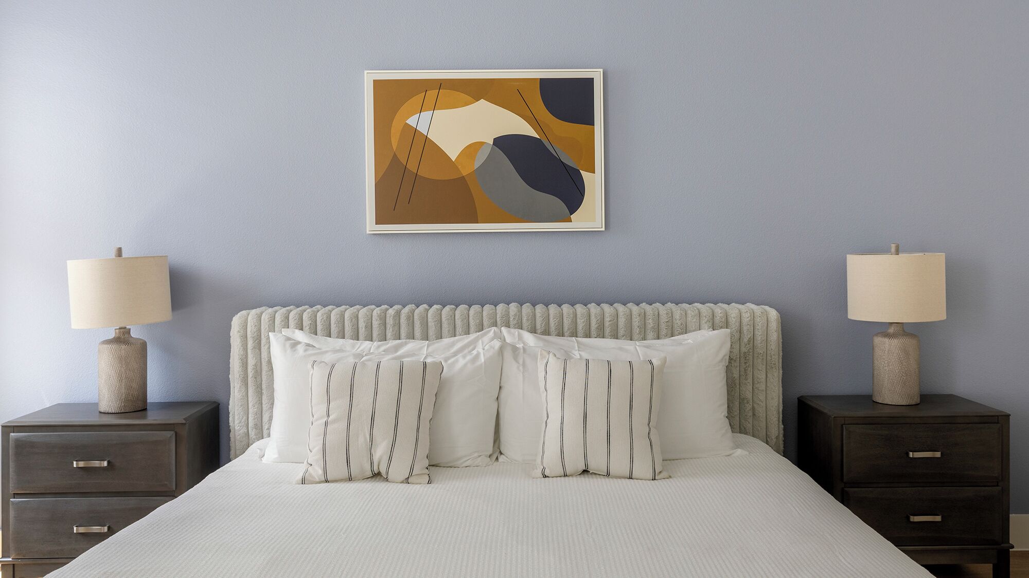 A bedroom with a bed, two nightstands, two lamps, and a framed abstract painting on the wall.