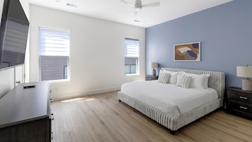 Un dormitorio con una cama grande, dos mesitas de noche, un buró, un ventilador de techo y dos ventanas con persianas.