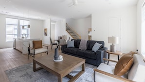 Uma sala de estar moderna com um sofá cinza escuro, uma mesa de centro de madeira e um espaço de jantar com mesa e cadeiras brancas.