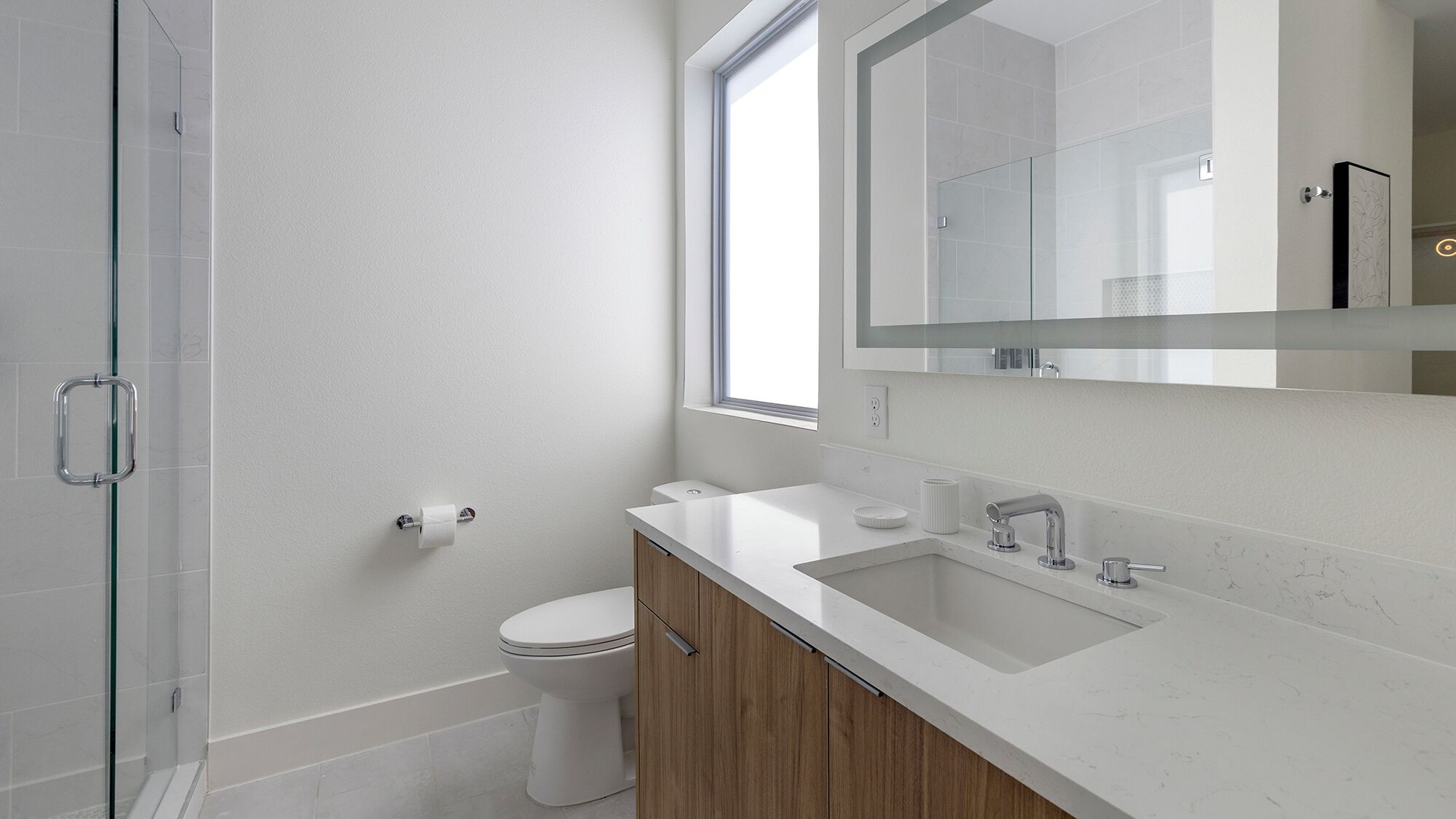 A modern bathroom with a glass shower enclosure, a toilet, a sink with a mirror above it, and a window providing natural light.