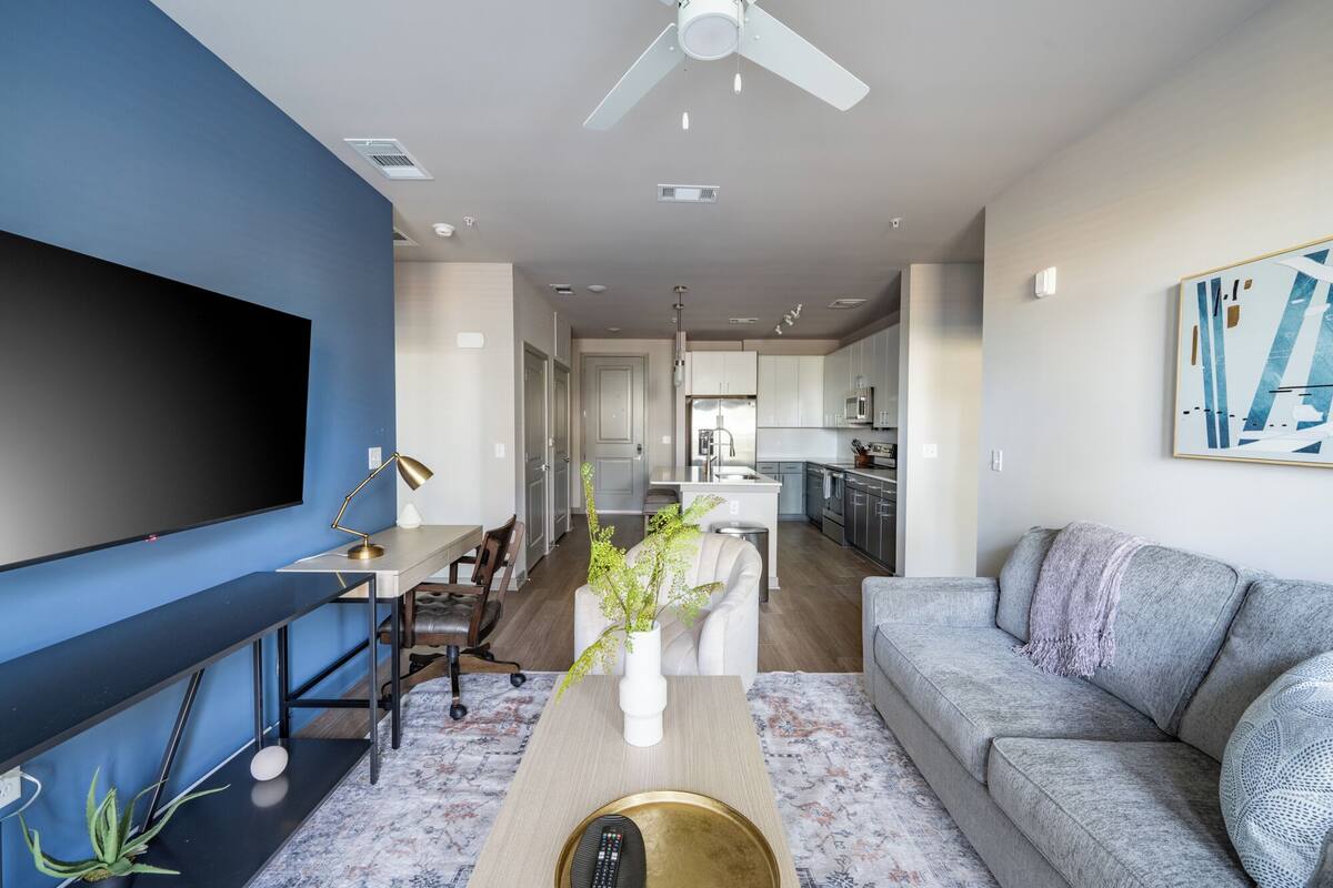 A modern living room with a flat-screen TV, a sofa, a coffee table, and a dining area with a chair and a lamp.