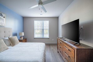 A bedroom with a bed, a dresser, a TV, a ceiling fan, and a window with blinds. - Landing Apartments - North Central Pensacola (Pensacola)