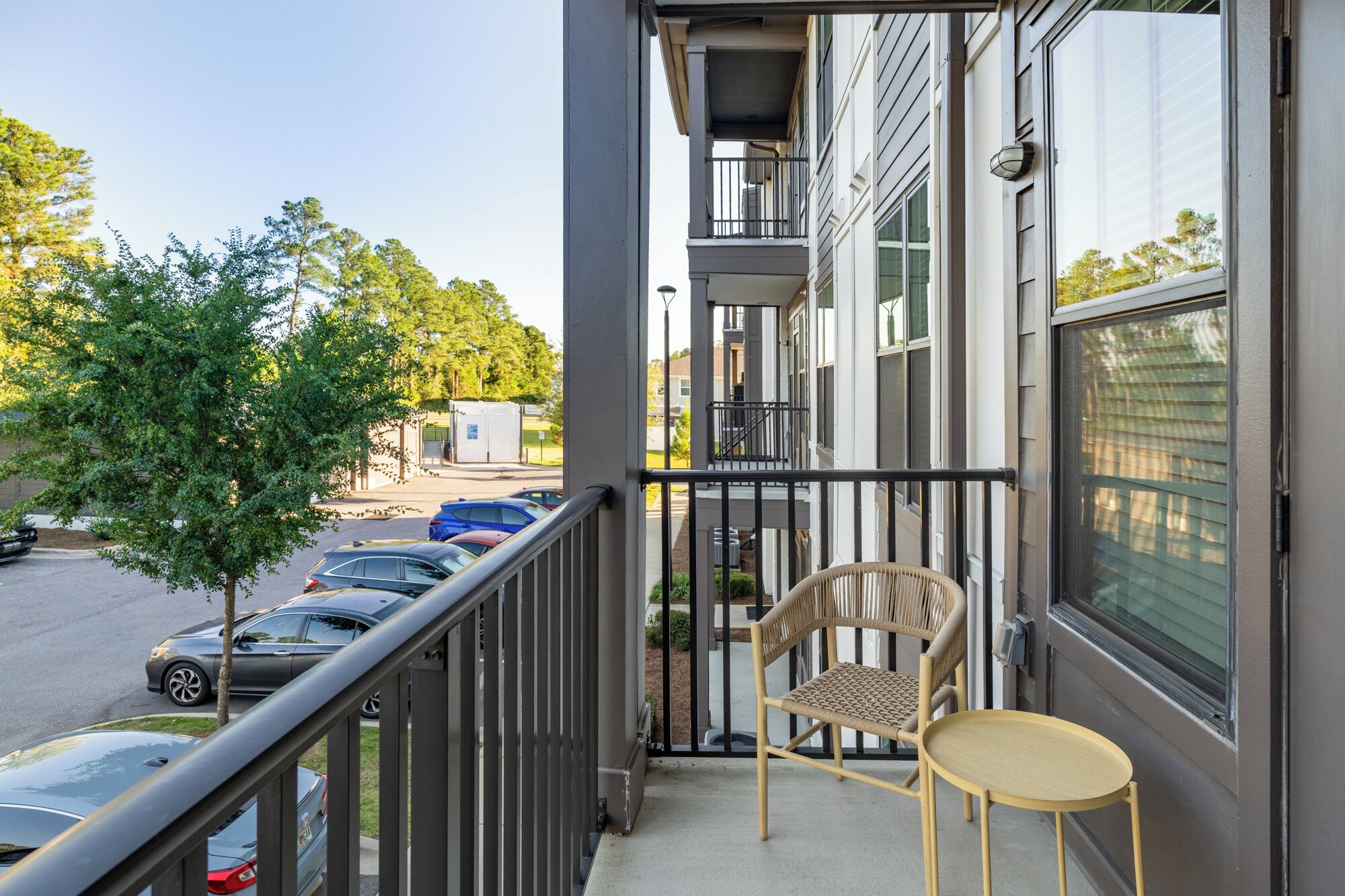 Un balcon avec une chaise et une table, donnant sur un parking et un immeuble doté de nombreuses fenêtres.