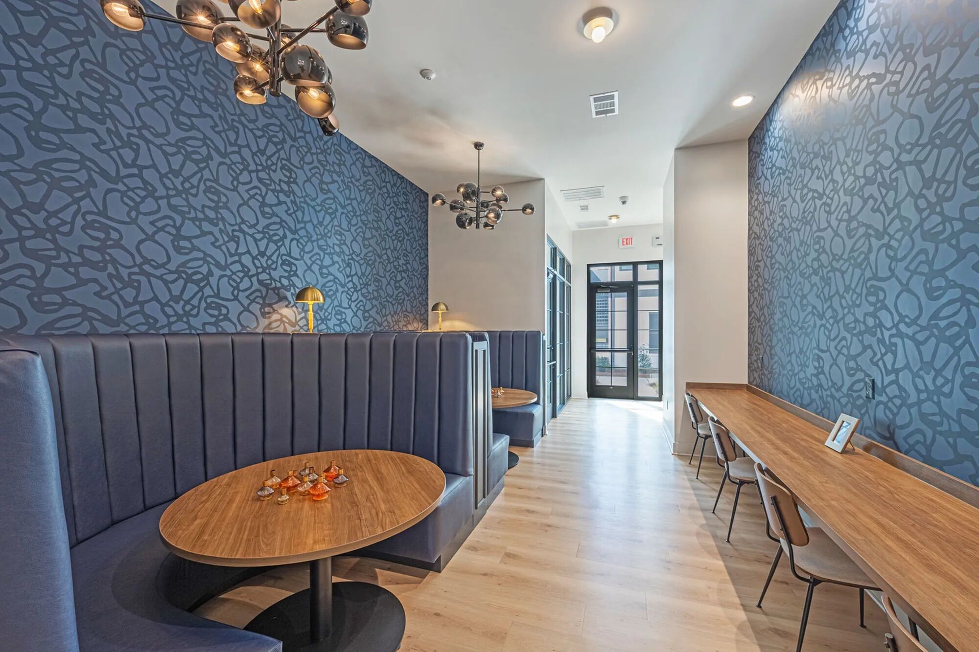 A modern restaurant with a long wooden table, cushioned seating, and patterned walls.