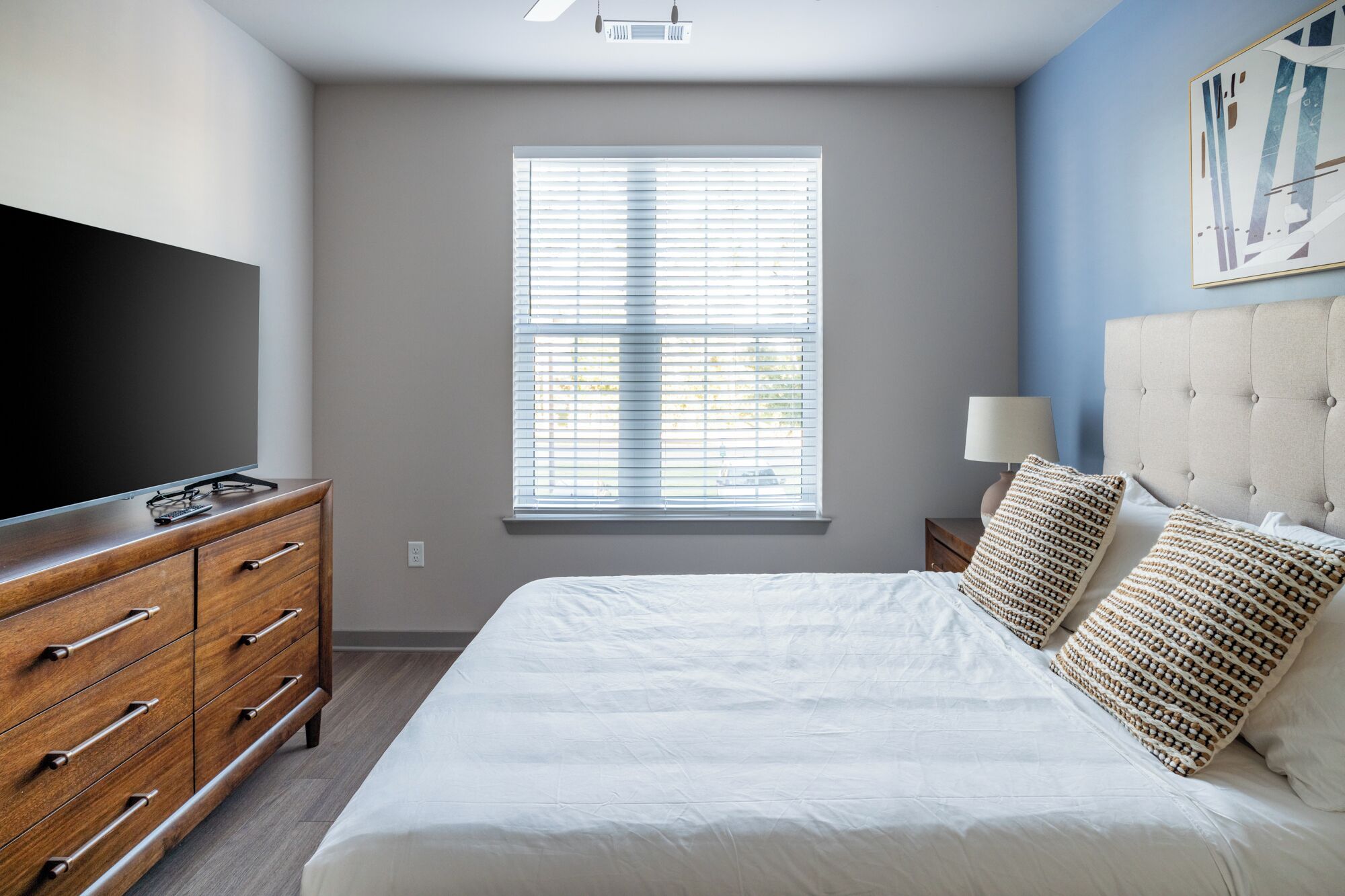 A bedroom with a bed, a TV on a wooden dresser, a window with blinds, and a nightstand with a lamp.