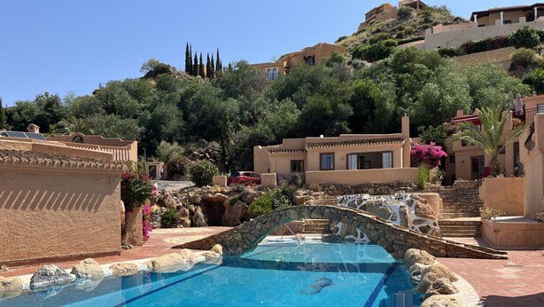 A pool with a stone bridge, surrounded by buildings with Mediterranean architecture, set against a hillside backdrop. - El Oasis - Boutique Mountain Retreat with Pool • 20 mins from Mojácar Beaches (AN)