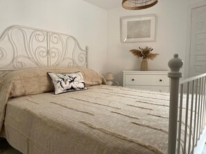 Een slaapkamer met een wit metalen hoofdbord, een beige sprei, een zwart-wit kussen, een witte nachtkastje met een vaas gedroogde bloemen, en een lijst met een afbeelding aan de muur.