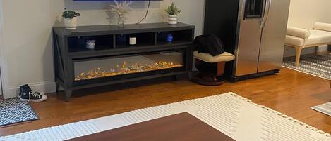 Een woonkamer met een flatscreen-tv waarop een nieuwsuitzending wordt vertoond, een donker houten salontafel, een zwarte entertainmentkast en een roestvrijstalen koelkast.
