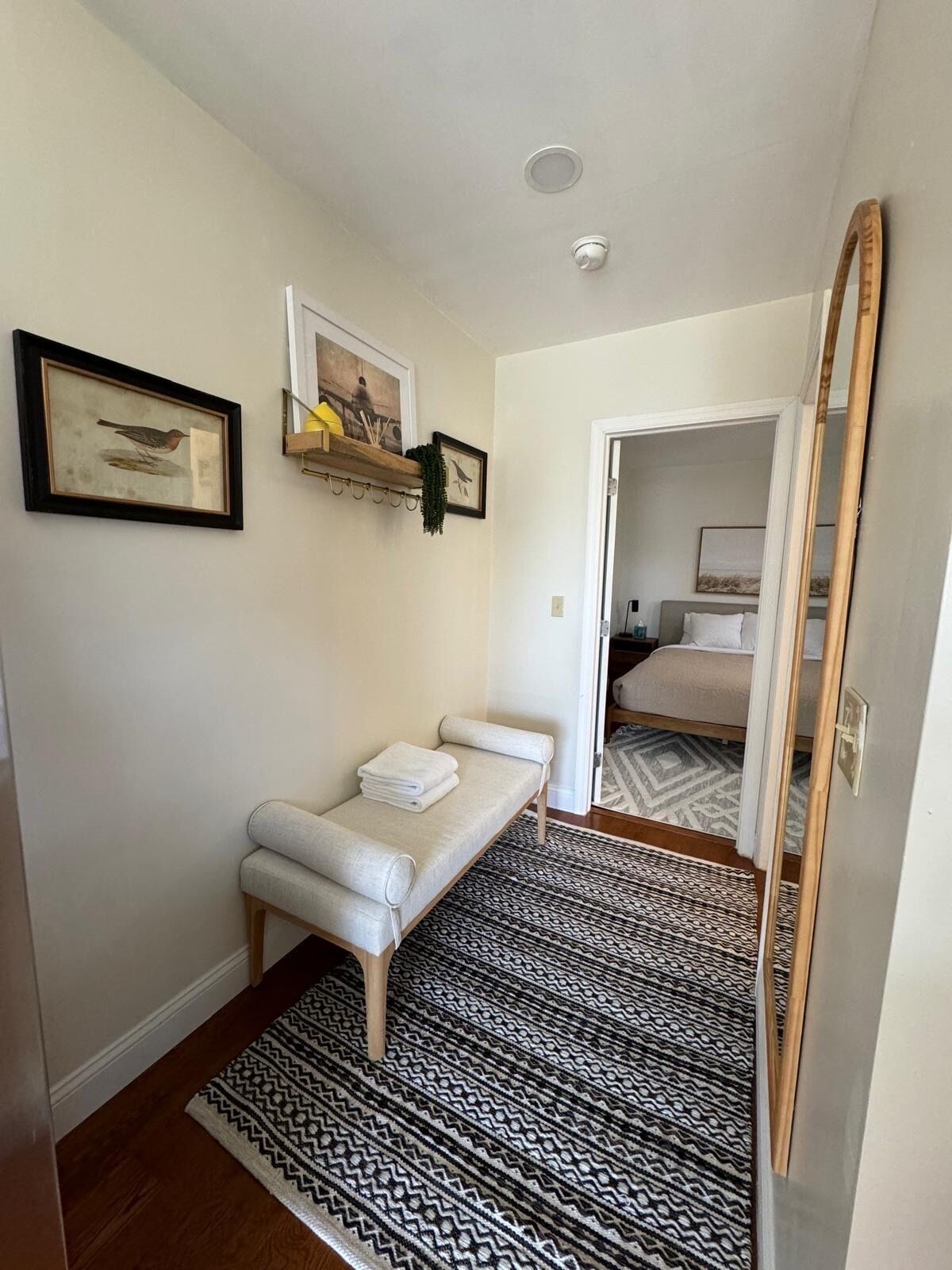 Une petite chambre à coucher, bien rangée, avec un lit simple, une table de chevet avec des serviettes, un tapis à motifs et un miroir.