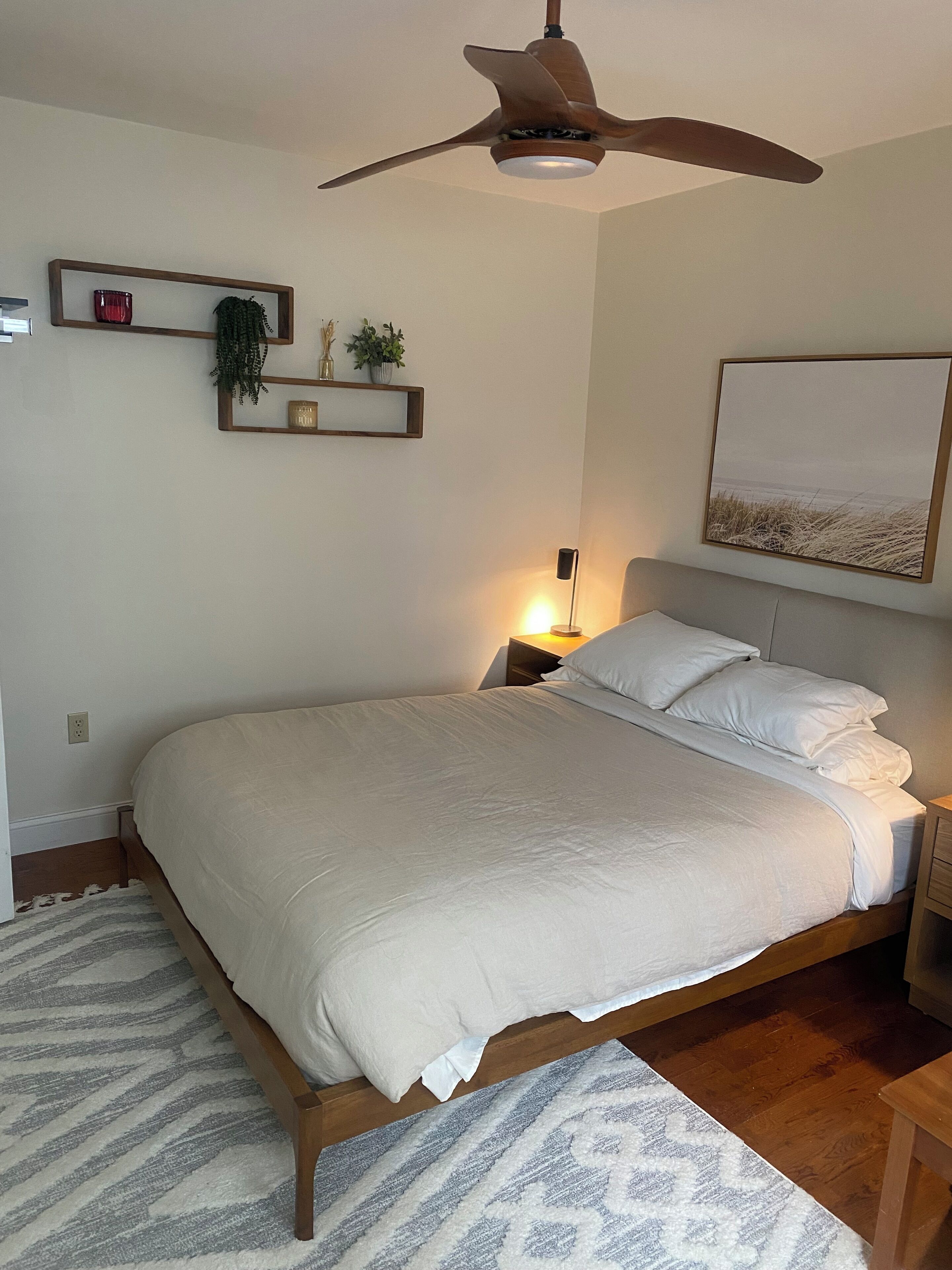 A bedroom with a large bed, a ceiling fan, a bedside lamp, a framed picture on the wall, and a small shelf with decorative items.