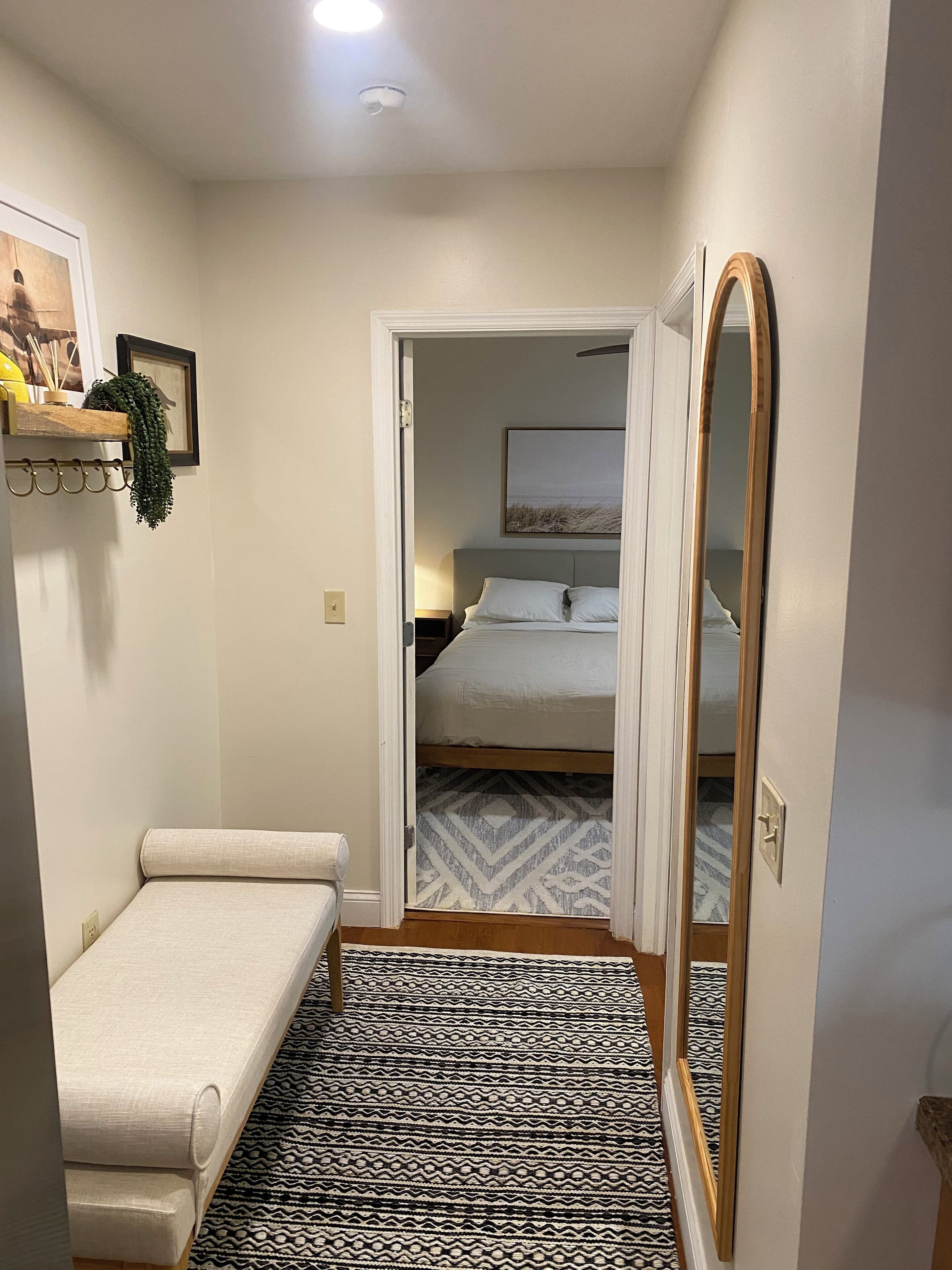 A narrow hallway leading to a bedroom with a bed, a bedside table, and a mirror.