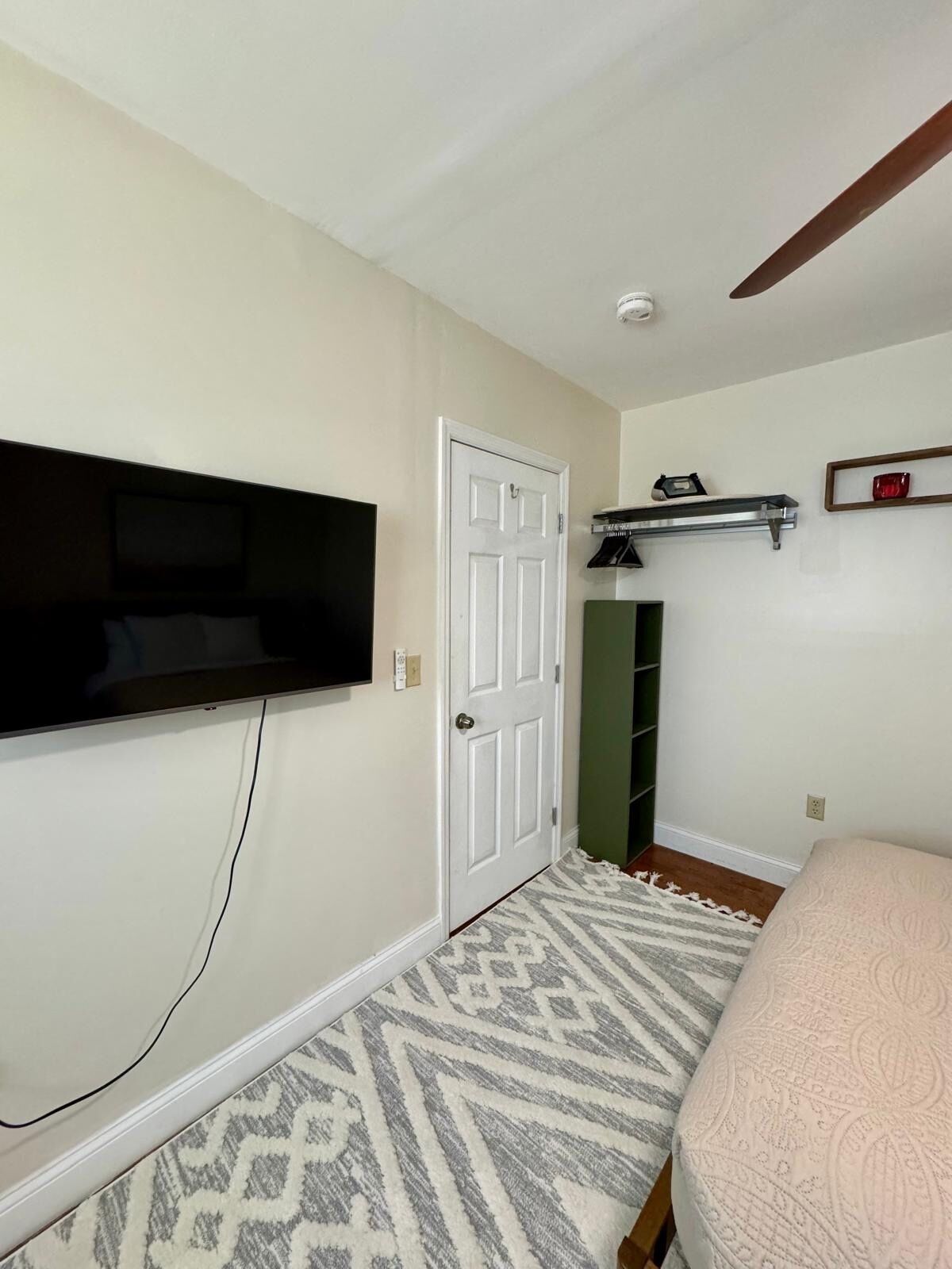 A bedroom with a bed, a television mounted on the wall, a door, and a shelf with items.