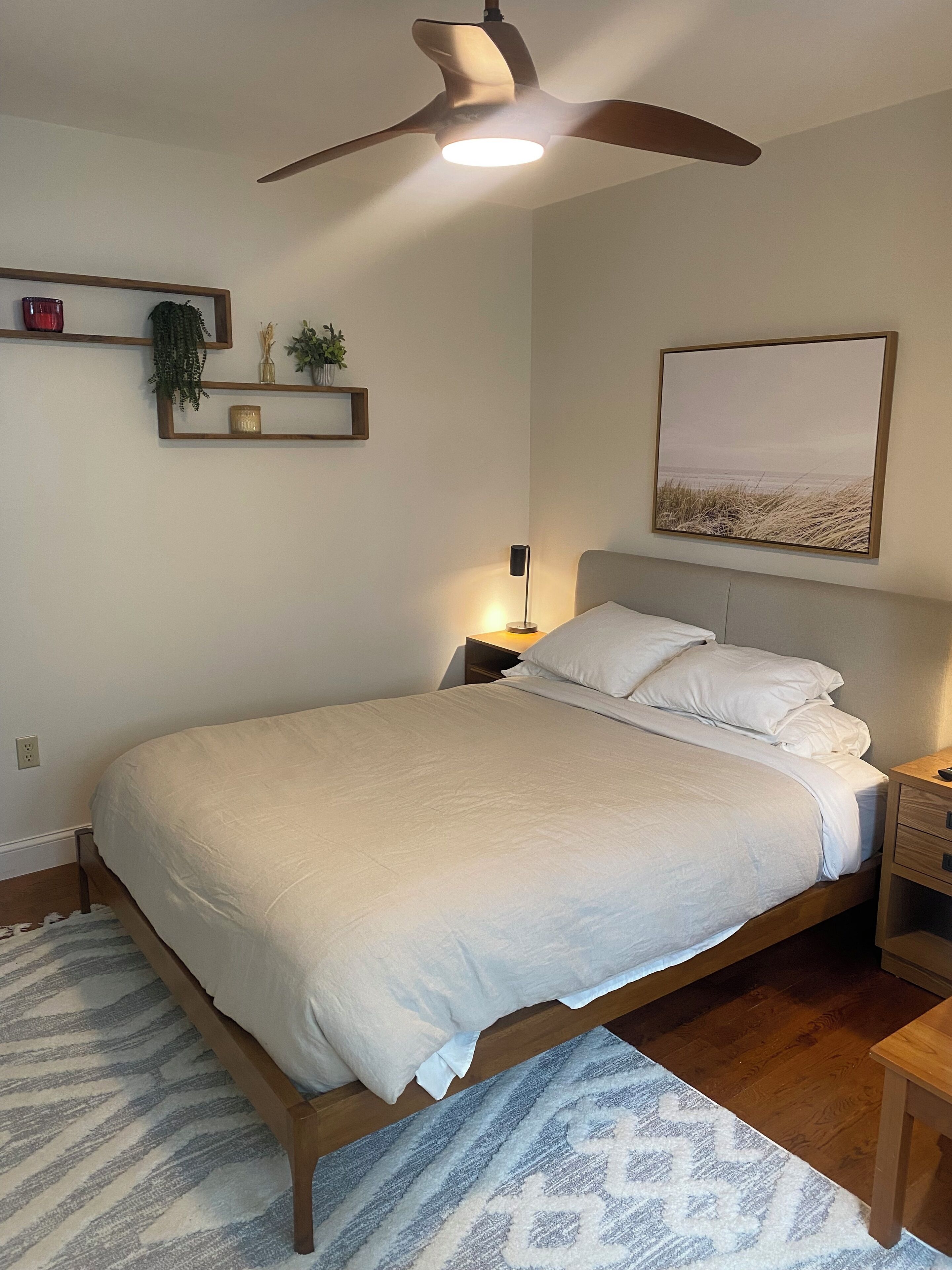 A bedroom with a bed, a ceiling fan, a bedside table, a nightstand, a lamp, a framed picture, and a small shelf with decorative items.