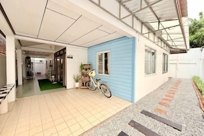 A covered outdoor area with a tiled floor, a blue wall, a bicycle, and a pathway leading to a door. - Living Room Udonthani (Udon Thani)