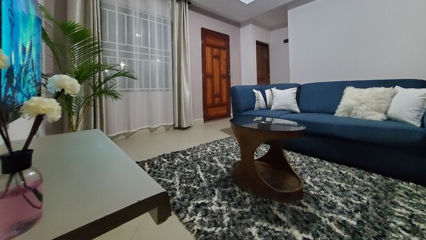 Un salon avec un canapé bleu, une table basse en verre, une table d’appoint blanche, un tapis à motifs et une grande fenêtre ornée de rideaux légers.