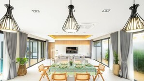 Un comedor moderno con una mesa grande dispuesta para seis personas, rodeada de sillas de madera. Hay tres lámparas colgantes sobre la mesa, y un televisor de pantalla plana montado en la pared detrás de la mesa.