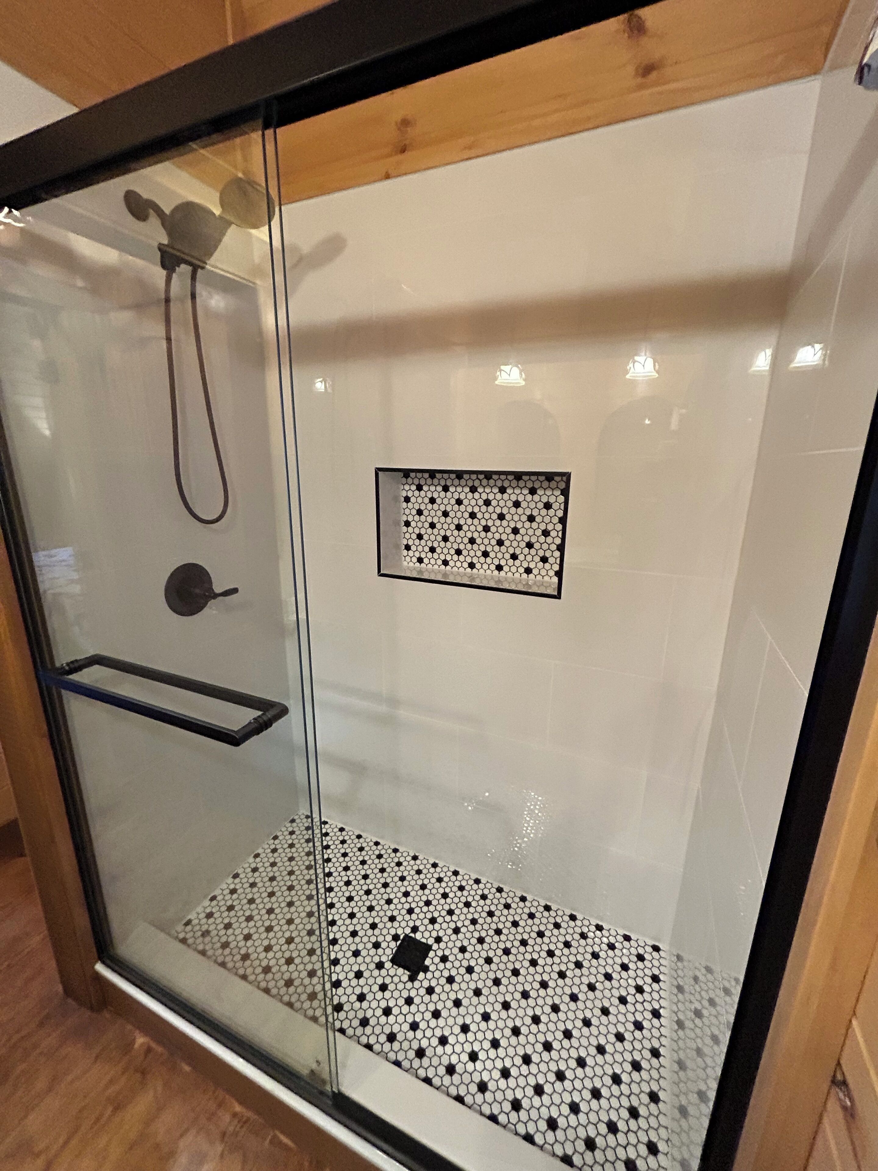 A glass-enclosed shower with a black and white tiled floor, a handheld showerhead, and a small rectangular vent.
